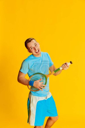 Photo pour Portrait de jeune homme en uniforme bleu jouant de la raquette de tennis comme de la guitare, posant isolé sur fond jaune. Amusant. Concept de sport, mode de vie fitness, soins du corps, santé, jeunesse, action. Publicité - image libre de droit