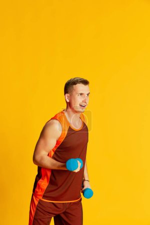 Téléchargez les photos : Portrait de jeune homme aux mains musclées en uniforme orange dressé avec des poids, posé isolé sur fond jaune. Concept de sport, mode de vie fitness, soins du corps, santé, jeunesse, action. Publicité - en image libre de droit
