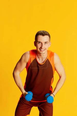 Téléchargez les photos : Portrait de jeune homme en uniforme orange, posant avec des équipements sportifs isolés sur fond jaune. Concept de sport, mode de vie fitness, soins du corps, santé, jeunesse, action. Publicité - en image libre de droit