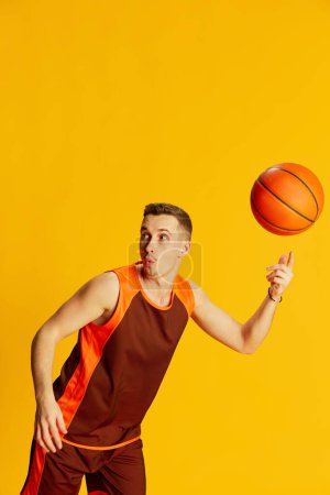 Téléchargez les photos : Portrait d'un jeune homme émotif en uniforme orange jouant au basket-ball, s'entraînant, tournant la balle sur un doigt isolé sur fond jaune. Concept de sport, mode de vie fitness, soins du corps, santé, action. Publicité - en image libre de droit