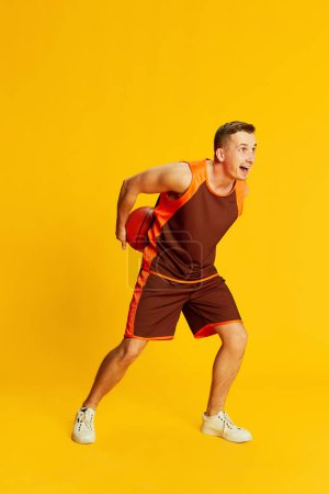 Téléchargez les photos : Portrait de jeune homme actif en uniforme orange, jouant au basket isolé sur fond jaune. Concept de sport, mode de vie fitness, soins du corps, santé, jeunesse, action. Publicité - en image libre de droit