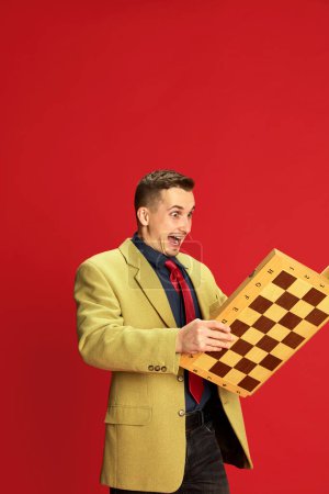 Photo for Portrait of young man in stylish jacket and glasses posing with chess board over red background. Happy and excited. Concept of emotions, business, occupation, hobby, lifestyle, fashion - Royalty Free Image
