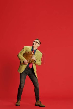Photo for Portrait of young happy man in stylish jacket, glasses and vintage briefcase posing over red background. Successful job interview. Concept of emotions, business, occupation, hobby, lifestyle, fashion - Royalty Free Image