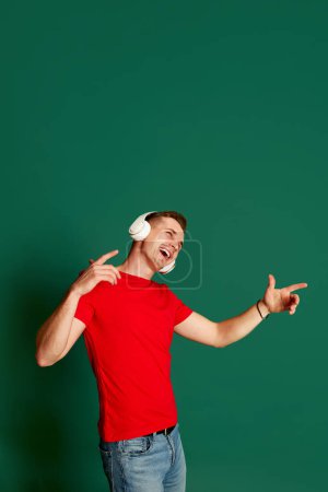 Photo for Portrait of young emotive man in casual clothes posing in headphones, singing isolated over green background. Concept of youth, lifestyle, music, casual fashion, emotions, facial expression. Ad - Royalty Free Image