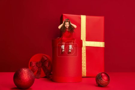 Photo for Cute little girl, child with curly hair sitting on giant gift boxed over red background. Happy New Year. Concept of holidays, celebration, presents, greetings. shopping, sales. Copy space for ad - Royalty Free Image