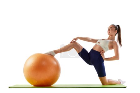 Photo for Portrait of young sportive woman training, doing stretching exercises with rubber fitness ball isolated over white background. Concept of sport, strength, body care, fitness, wellbeing, health. Ad - Royalty Free Image