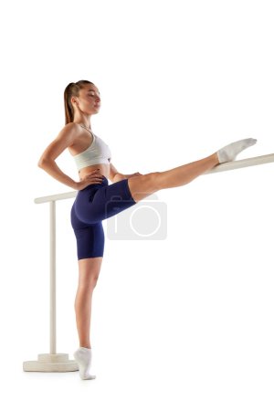 Photo for Full-length portrait of young sportive woman training, doing stretching exercises on ballet barre isolated over white background. Concept of sport, strength, body care, fitness, wellbeing, health. Ad - Royalty Free Image