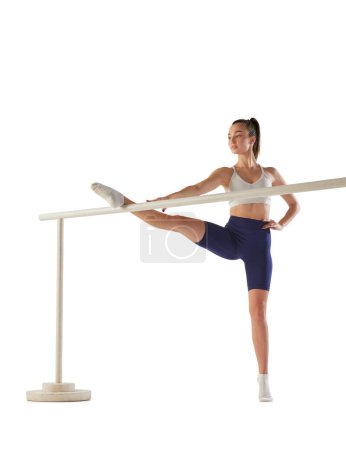 Photo for Portrait of young sportive woman training, doing stretching exercises on ballet barre isolated over white background. Dancer. Concept of sport, strength, body care, fitness, wellbeing, health. Ad - Royalty Free Image
