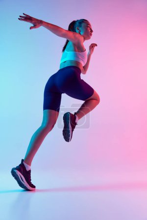 Foto de Vista inferior del entrenamiento de la mujer joven deportiva, corriendo aislado sobre el fondo azul degradado de color rosa en luz de neón. Concepto de deporte, fuerza, cuidado corporal, fitness, bienestar, salud. Anuncio - Imagen libre de derechos