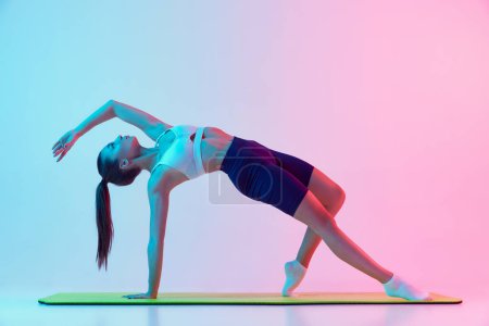Photo for Portrait of young sportive woman training, doing stretching exercises isolated over gradient blue pink background in neon light. Concept of sport, strength, body care, fitness, wellbeing, health. Ad - Royalty Free Image
