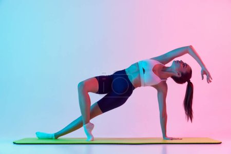 Photo for Portrait of young sportive woman training, standing on side plank isolated over gradient blue pink background in neon light. Concept of sport, strength, body care, fitness, wellbeing, health. Ad - Royalty Free Image