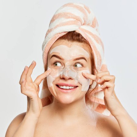 Photo for Portrait of young, beautiful, red-haired woman applying face clay mask after shower isolated on white background. Concept of beauty, face and skin care, cosmetology and natural cosmetics, spa - Royalty Free Image