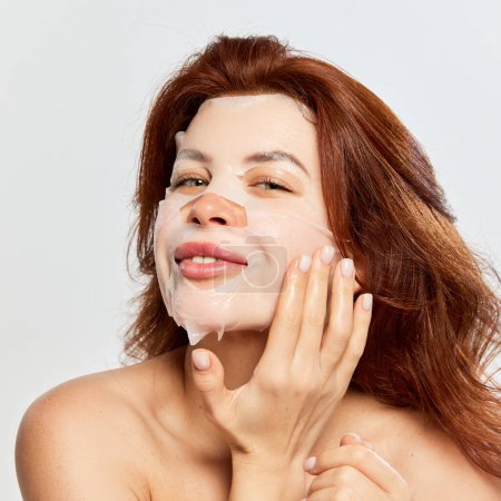 Photo for Beautiful, red-haired woman taking care after skin with moisturizing and revitalizing mask isolated on white background. Concept of beauty, face and skin care, cosmetology and natural cosmetics, spa - Royalty Free Image