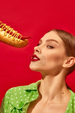 Foto de Mujer joven con maquillaje de moda comiendo hot dog picante con mostaza y chile sobre fondo rojo vivo. Pimienta en lugar de salchicha. Fotografía de arte pop de comida. Colores complementarios. Copiar espacio para anuncios, texto - Imagen libre de derechos