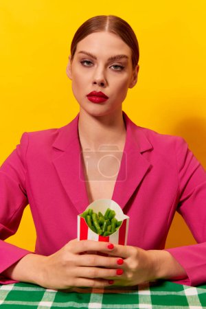 Foto de Mujer joven, seria y sin emociones, con una chaqueta rosa brillante sosteniendo judías verdes sobre un fondo amarillo. Dieta saludable. Fotografía de arte pop de comida. Colores complementarios. Copiar espacio para anuncios, texto - Imagen libre de derechos