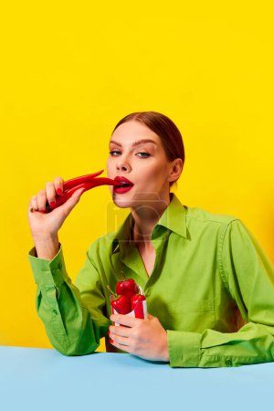 Foto de Hermosa mujer joven en chaqueta verde vintage comiendo chile rojo sobre fondo amarillo. Amante de la comida picante. Fotografía de arte pop de comida. Colores complementarios. Copiar espacio para anuncios, texto - Imagen libre de derechos