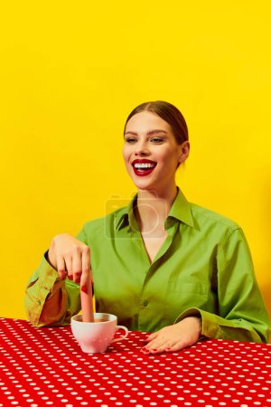 Foto de Mujer joven sonriente ropa vintage sumergiendo salchicha en la taza de café, capuchino sobre mantel rojo sobre fondo amarillo. Fotografía de arte pop de comida. Colores complementarios. Copiar espacio para anuncios, texto - Imagen libre de derechos