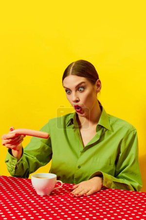 Téléchargez les photos : Jeune femme émotionnelle vêtements vintage manger saucisse avec café, cappuccino sur nappe rouge sur fond jaune. Photographie pop art culinaire. Couleurs complémentaires. Espace de copie pour annonce, texte - en image libre de droit