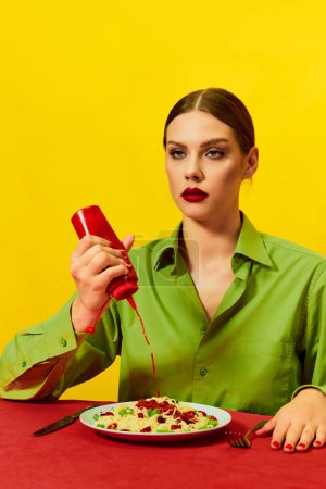 Foto de Chica joven con la cara sin emociones verter ketchup en el plato con espaguetis en mantel rojo sobre fondo amarillo. Picante. Fotografía de arte pop de comida. Colores complementarios. Copiar espacio para anuncios, texto - Imagen libre de derechos