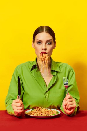 Photo for Young girl eating spaghetti, noodles sticking out of the mouth over yellow background. Dinner time. Emotionless, poker face. Food pop art photography. Complementary colors. Copy space for ad, text - Royalty Free Image