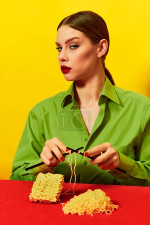 Foto de Mujer con la cara sin emociones tejiendo fideos instantáneos en la mesa azul sobre un mantel rojo vivo sobre fondo amarillo. Fotografía de arte pop de comida. Colores complementarios. Copiar espacio para anuncios, texto - Imagen libre de derechos