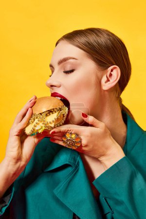 Foto de Joven hermosa mujer en abrigo verde comiendo, mordiendo hamburguesa con collares sobre fondo amarillo. Comida de lujo. Fotografía de arte pop de comida. Colores complementarios. Copiar espacio para anuncios, texto - Imagen libre de derechos