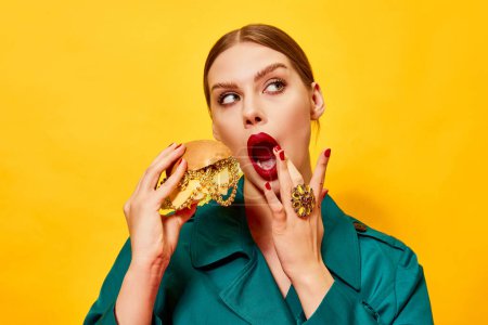 Foto de Joven hermosa mujer en abrigo verde con lápiz labial rojo comiendo hamburguesa con collares sobre fondo amarillo. Fotografía de arte pop de comida. Colores complementarios. Copiar espacio para anuncios, texto - Imagen libre de derechos