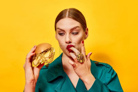 Foto de Joven hermosa mujer en abrigo verde con lápiz labial rojo comiendo hamburguesa con collares, lamiendo los dedos sobre fondo amarillo. Fotografía de arte pop de comida. Colores complementarios. Copiar espacio para anuncio - Imagen libre de derechos