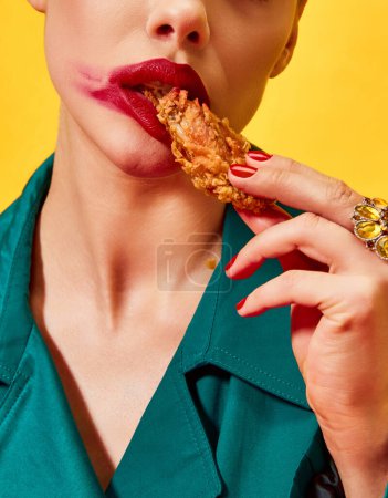 Téléchargez les photos : Image recadrée d'une femme en manteau vert avec du rouge à lèvres, mangeant du poulet frit, des pépites sur fond jaune. Restauration rapide. Photographie pop art culinaire. Couleurs complémentaires. Espace de copie pour annonce, texte - en image libre de droit