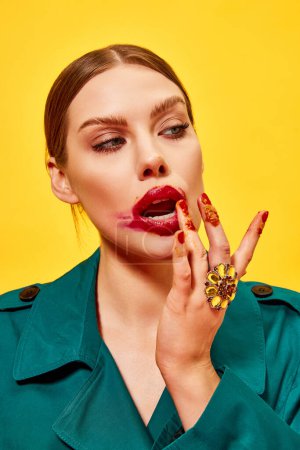 Photo for Young beautiful woman in green coat with smudged red lipstick licking fingers after eating nuggets over yellow background. Food pop art photography. Complementary colors. Copy space for ad, text - Royalty Free Image
