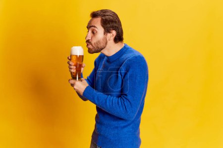 Téléchargez les photos : Portrait d'un homme émotif en pull bleu posant avec une tasse de bière sur fond jaune. Souffler sur la mousse. Goût traditionnel. Concept d'émotions, dégustation de bière, style de vie, expression faciale, Oktoberfest - en image libre de droit
