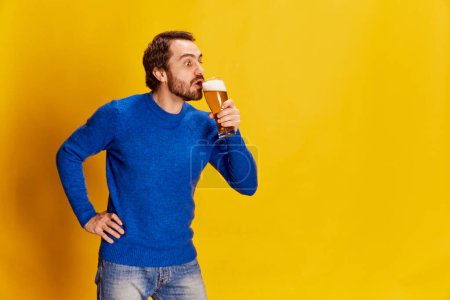 Photo for Portrait of mature man in blue sweater posing with beer glass, sipping lager beer over yellow background. Weekends. Concept of emotions, beer degustation, lifestyle, facial expression, Oktoberfest - Royalty Free Image