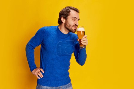 Foto de Hombre emotivo en jersey azul posando con jarra de cerveza, bebiendo cerveza espumosa lager con placer aislado sobre fondo amarillo. Emociones, degustación de cerveza, estilo de vida, expresión facial, concepto Oktoberfest - Imagen libre de derechos
