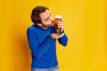 Téléchargez les photos : Portrait d'homme en pull bleu posant avec une tasse de bière, embrassant verre avec boisson mousseuse sur fond jaune. Goût de houblon. Concept d'émotions, dégustation de bière, style de vie, expression faciale, Oktoberfest - en image libre de droit