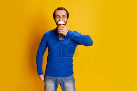 Photo for Portrait of emotive man in blue sweater posing with drink glass, cheerfully sipping beer isolated on yellow background. Concept of emotions, beer degustation, lifestyle, facial expression, Oktoberfest - Royalty Free Image