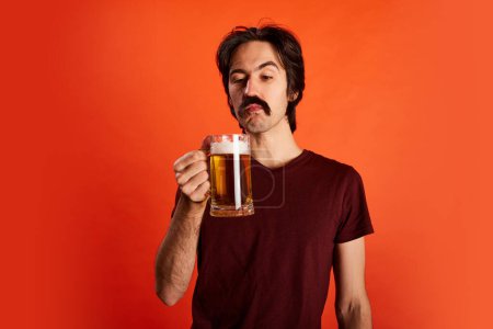 Foto de Retrato de un hombre emotivo con bigote posando con un vaso de cerveza fresca, espumosa y lager aislada sobre fondo rojo. Probando un nuevo sabor. Concepto de emociones, degustación de cerveza, expresión facial, Oktoberfest - Imagen libre de derechos