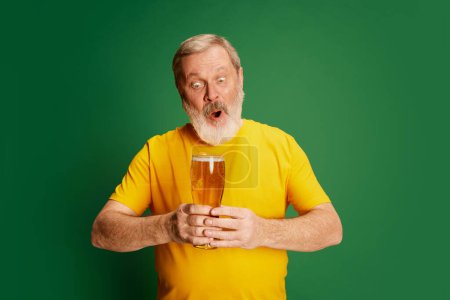 Téléchargez les photos : Portrait d'homme âgé en T-shirt jaune posant émotionnellement avec du verre à bière isolé sur fond vert. Délicieux. Concept d'émotions, dégustation de bière, style de vie, expression faciale, Oktoberfest - en image libre de droit