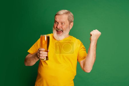Téléchargez les photos : Portrait d'homme âgé en T-shirt jaune posant émotionnellement avec de la bière isolée sur fond vert. Fan de football. Concept d'émotions, dégustation de bière, style de vie, expression faciale, Oktoberfest - en image libre de droit