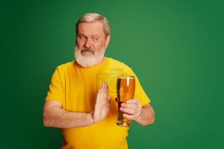 Téléchargez les photos : Portrait d'homme âgé en T-shirt jaune posant avec de la bière isolée sur fond vert. Rejeter les boissons alcoolisées. Concept d'émotions, dégustation de bière, style de vie, expression faciale, Oktoberfest - en image libre de droit