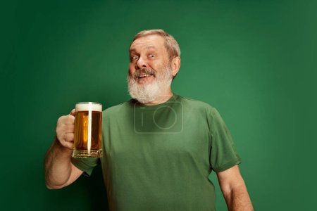 Foto de Retrato de hombre mayor feliz y emocionado en camiseta posando con cerveza aislada sobre fondo verde. Hora de la fiesta. Concepto de emociones, degustación de cerveza, estilo de vida, expresión facial, Oktoberfest - Imagen libre de derechos