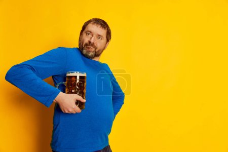 Téléchargez les photos : Portrait d'un homme émotif et drôle en pull bleu posant avec une tasse de bière mousseuse isolée sur fond jaune. Concept d'émotions, dégustation de bière, style de vie, expression faciale, Oktoberfest. Affiche, annonce - en image libre de droit