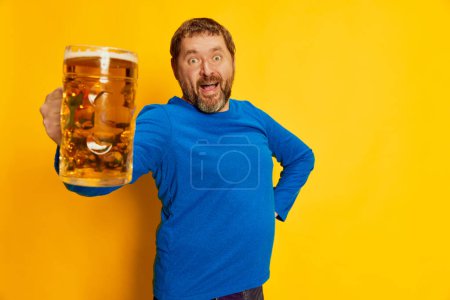 Téléchargez les photos : Portrait d'homme émotif en pull bleu posant joyeusement avec une tasse de bière mousseuse isolée sur fond jaune. Excité. Concept d'émotions, dégustation de bière, style de vie, expression faciale, Oktoberfest - en image libre de droit