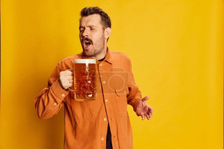 Téléchargez les photos : Beau homme en chemise orange posant avec verre à bière mousseux lager isolé sur fond jaune. Ça a l'air heureux et excité. Concept d'émotions, dégustation de bière, style de vie, expression faciale, Oktoberfest - en image libre de droit