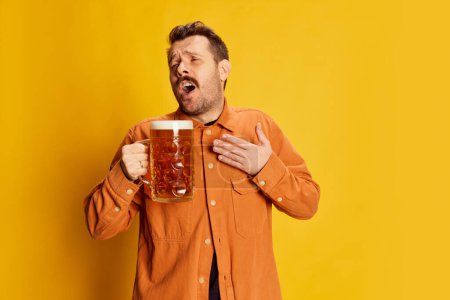 Foto de Retrato de hombre guapo emotivo en camisa naranja posando con vidrio de cerveza espumoso lager aislado sobre fondo amarillo. Bebida refrescante. Concepto de emociones, degustación de cerveza, estilo de vida, Oktoberfest - Imagen libre de derechos