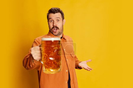 Foto de Invitación. Hombre guapo emotivo en camisa naranja posando con vidrio de cerveza espumoso lager aislado sobre fondo amarillo. Concepto de emociones, fiesta de la cerveza, estilo de vida, expresión facial, Oktoberfest - Imagen libre de derechos