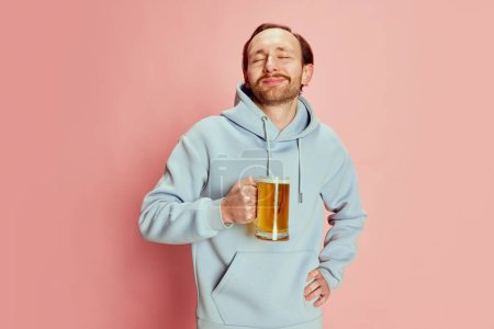 Photo for Portrait of young man in casual hoodie posing with lager foamy beer mug isolated over pink background. Happy and delightful. Concept of emotions, taste, lifestyle, facial expression, Oktoberfest - Royalty Free Image