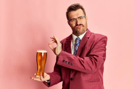 Foto de Retrato de hombre emotivo con estilo en un traje posando con un vaso de cerveza lager aislado sobre fondo rosa. El sabor perfecto de la cerveza. Concepto de emociones, gusto, estilo de vida, expresión facial, Oktoberfest - Imagen libre de derechos