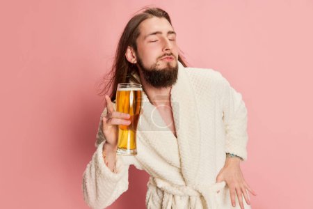 Foto de Retrato de hombre divertido emotivo en albornoz posando con un vaso de cerveza lager aislado sobre fondo rosa. Me siento bien. Concepto de emociones, gusto, estilo de vida, expresión facial, Oktoberfest - Imagen libre de derechos