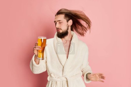 Téléchargez les photos : Portrait d'homme drôle émotif en peignoir posant avec tasse de bière lager isolé sur fond rose. Préparation de la fête. Concept d'émotions, goût, style de vie, expression faciale, Oktoberfest - en image libre de droit