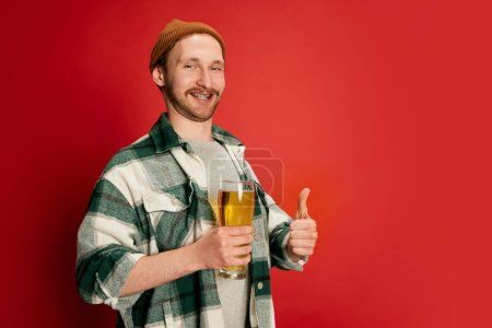 Téléchargez les photos : Portrait de jeune homme en chemise à carreaux décontractée posant avec du verre à bière isolé sur fond rouge. Elle a l'air heureuse. Goût parfait. Concept d'émotions, goût, style de vie, expression faciale, Oktoberfest - en image libre de droit